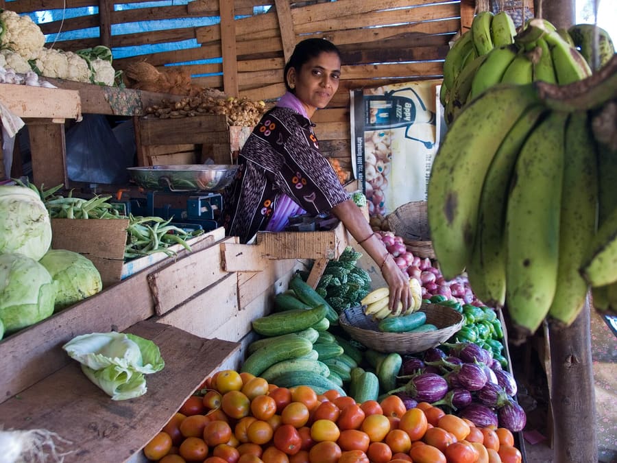 A woman arranging vegetables at a market stall, with bananas hanging in front